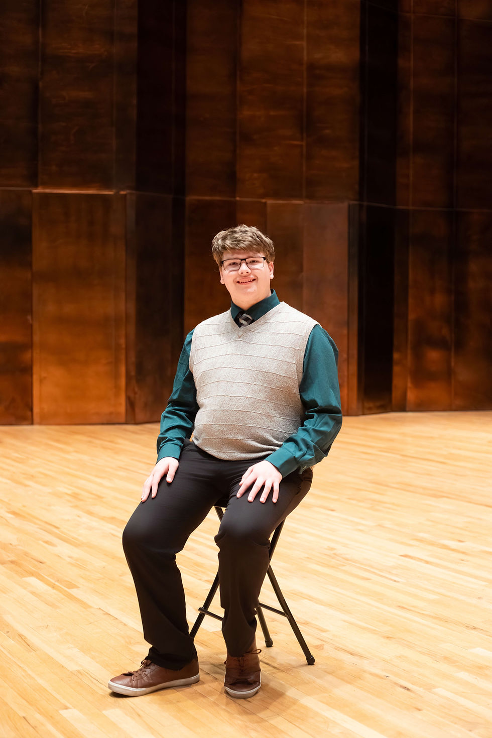 Young person smiling, sitting on a stool in a wooden-floored auditorium with brown panel walls, wearing a gray vest and teal shirt. Charleston High School senior photo Doudna Fine Arts Center.
