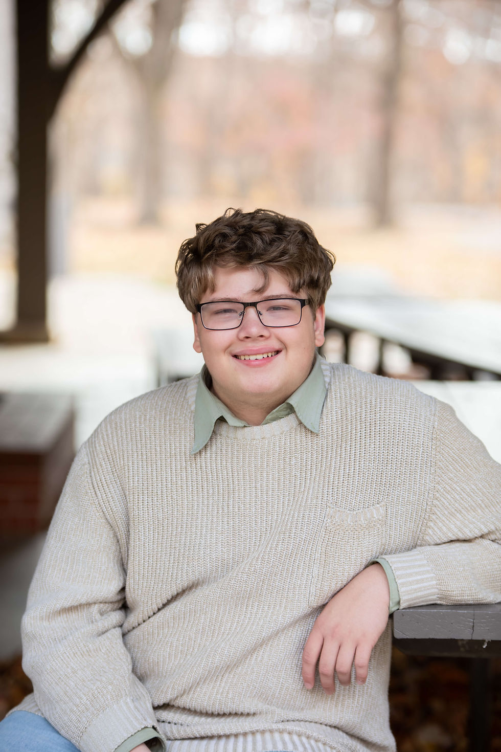Smiling person in glasses wearing a beige sweater, seated outdoors with autumn trees in the blurry background, exuding a warm, relaxed mood. Charleston High School senior photo Doudna Fine Arts Center.