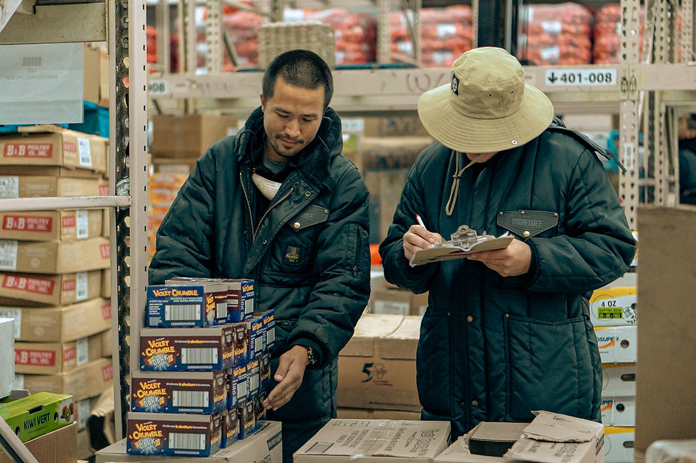 Two PDC Wholesale team members conducting inventory in a warehouse, reviewing products and taking notes on a clipboard beside stacked boxes of Violet Crumble candy.