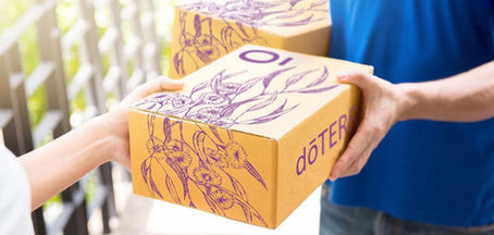 Hands exchange a brown box with purple floral designs and "doTERRA" text. Background shows sunlight on a blurred fence.