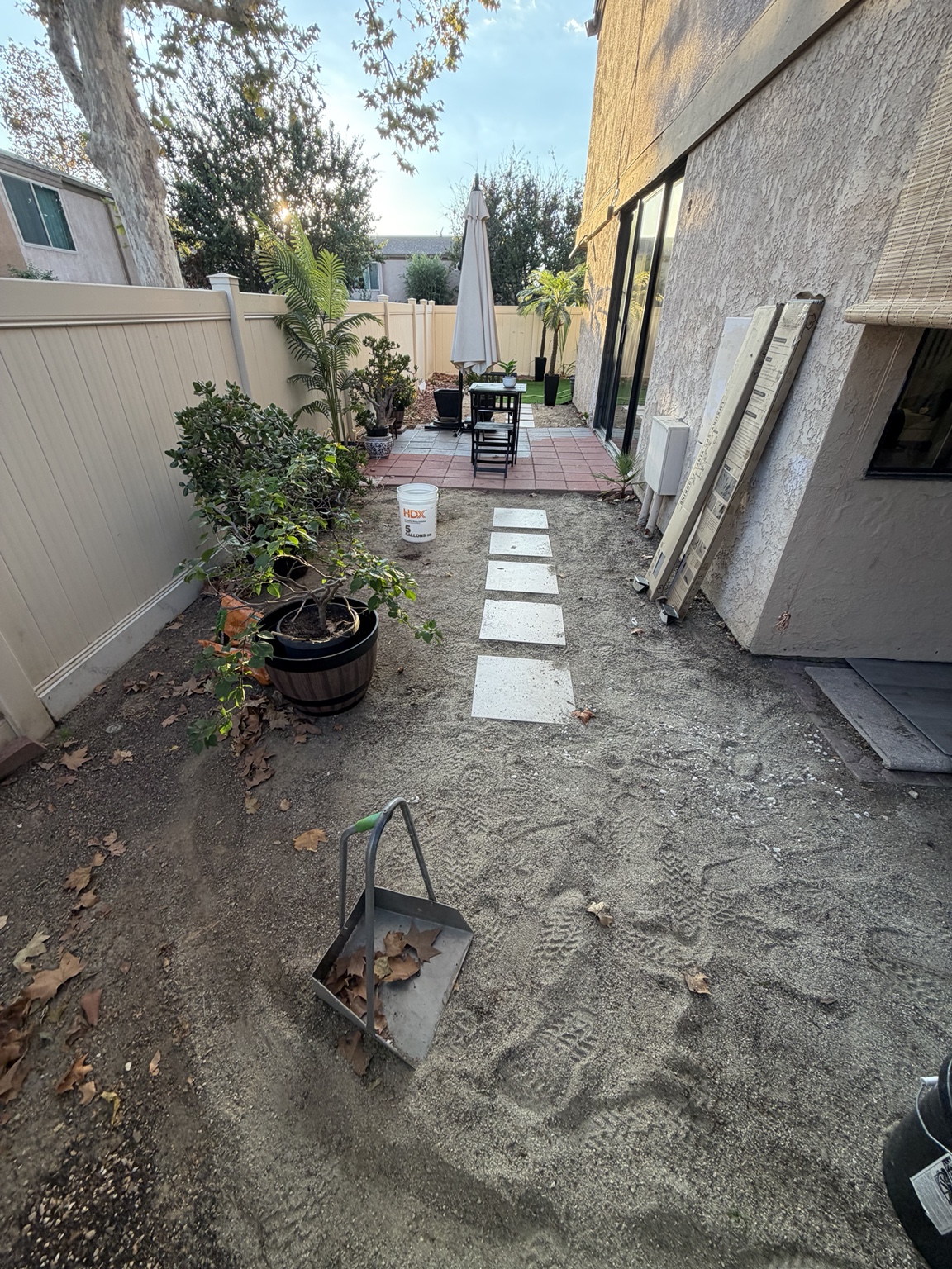 Original side-yard photo with bare ground, square stepping stones, potted plants and a vinyl fence