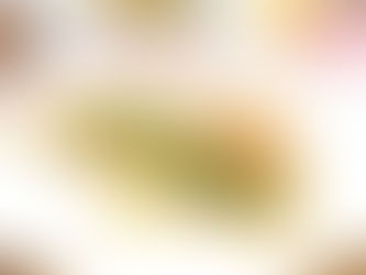 Slice of pastry with spinach and cheese filling on a white plate. Topped with sesame seeds. Background shows blurred bowls and soft lighting.