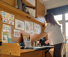 Lucy arranging artwork and paintbrushes on desk by watercolor paintings.