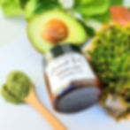 un barattolo di maschera viso olio di avocado e matcha, con mezzo avocado, polvere di matcha su un cucchiaio e muschio verde