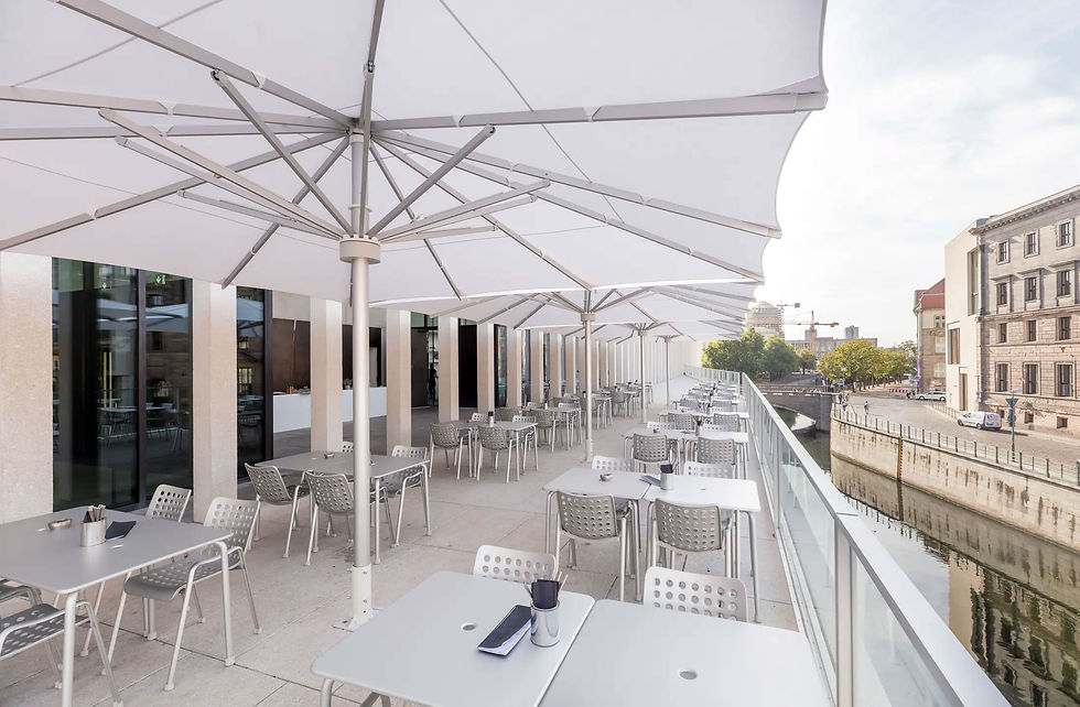 Mehrere weiße Großschirme in einer Reihe über einer langen Terrasse am Wasser, Tische und Stühle darunter