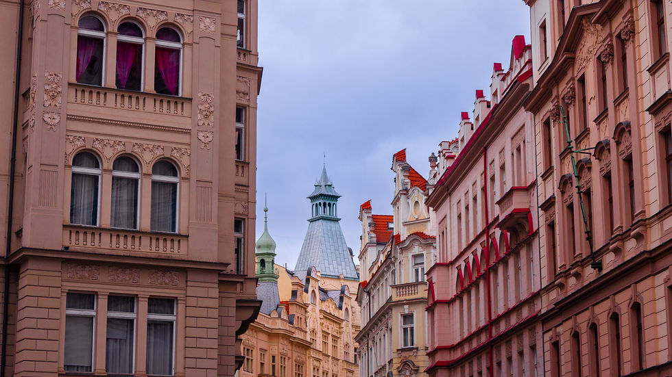 Ornate pastel buildings line a narrow street, with red roofs and towers under a cloudy sky, no people visible.