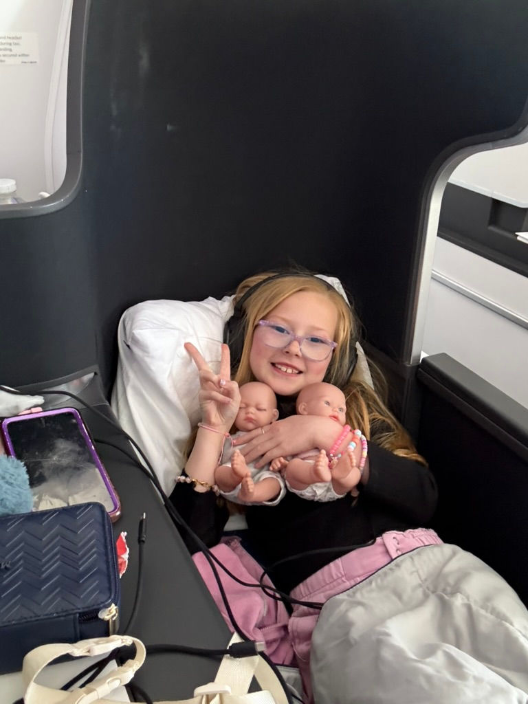 Girl smiling on a plane seat, holding two dolls, wearing glasses and headphones, showing a peace sign. Pink and black outfit, relaxed mood.