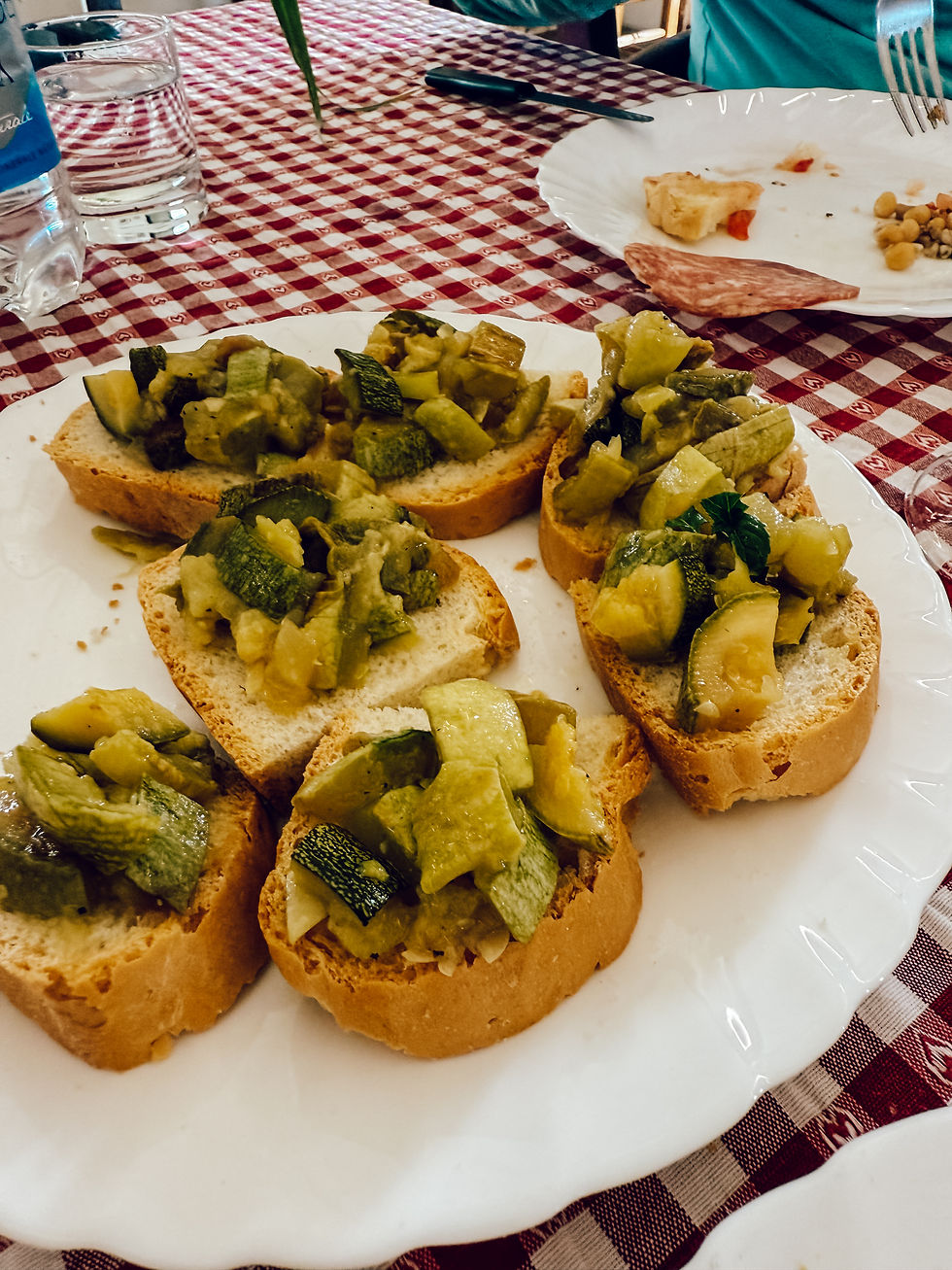 Bruschetta with green vegetable topping on white plate, red checkered tablecloth, and a glass of water nearby. Casual dining mood.