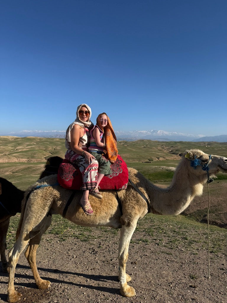 Two smiling women ride a camel across a grassy plain under a clear blue sky with distant mountains.