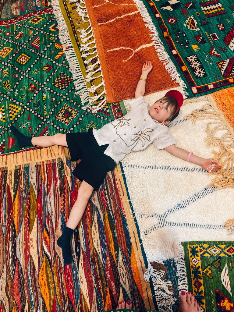 Child in shorts and shirt lies on colorful patterned rugs, looking content. Rugs feature vibrant greens, oranges, and reds.