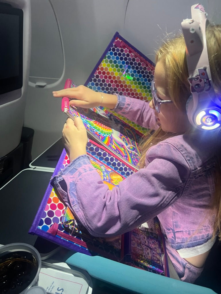 Child wearing headphones colors in a rainbow-themed book on an airplane. Bright colors and patterns, with soda on the tray table.