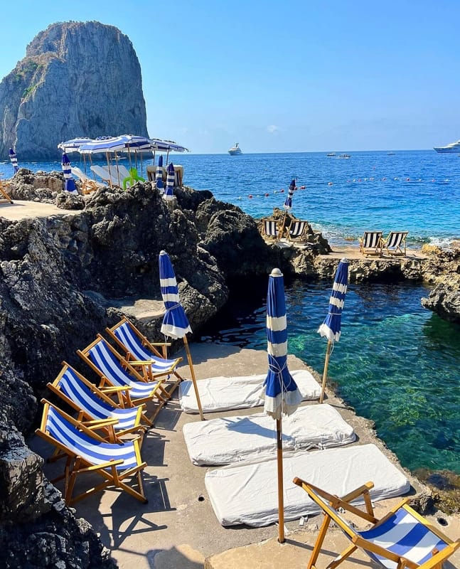 Capri beach club scene with striped umbrellas, rocky coastline, vivid blue water, Faraglioni energy.