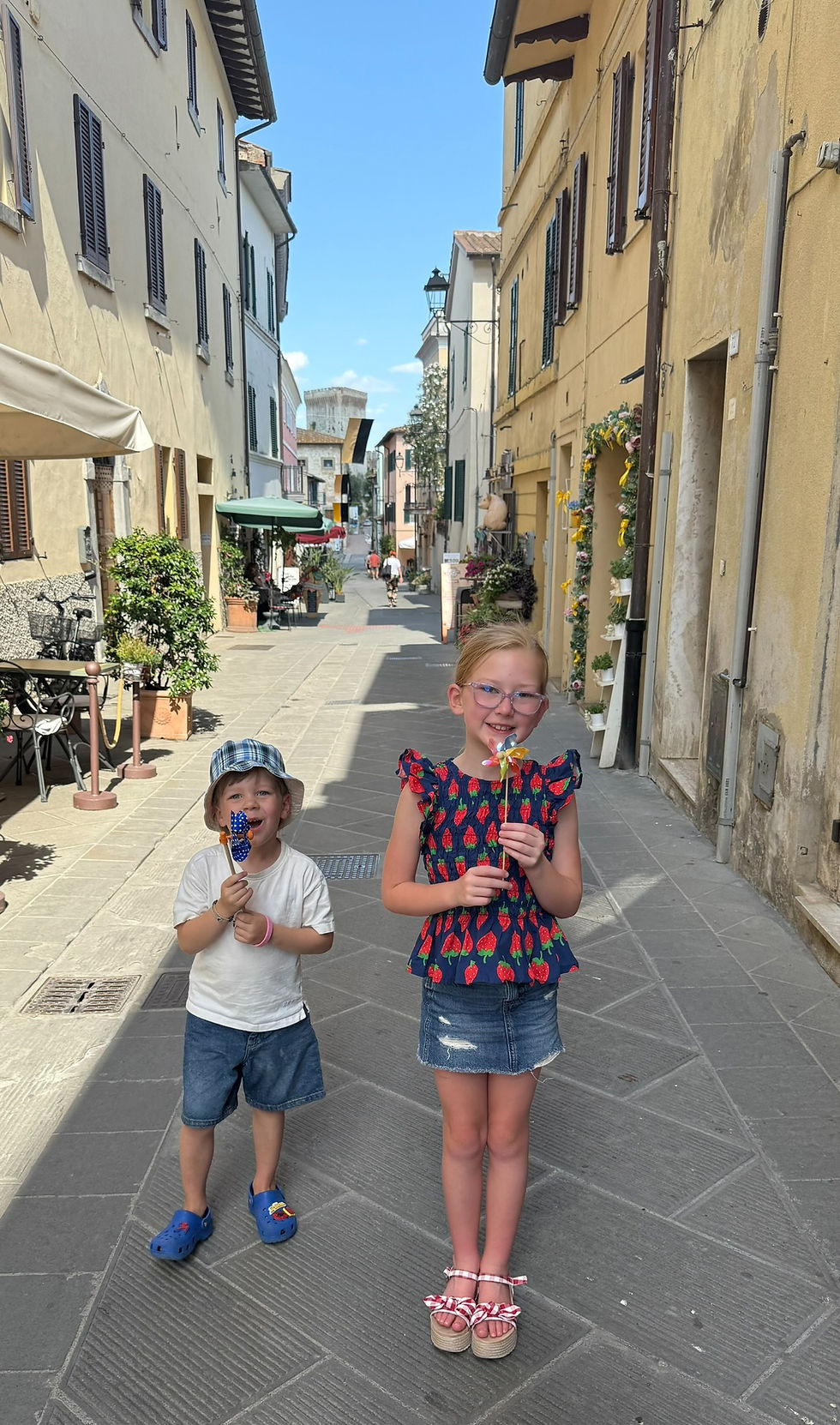 Two children smiling and holding pinwheels in a quaint street with pastel buildings and flower decorations on the walls under a clear sky.