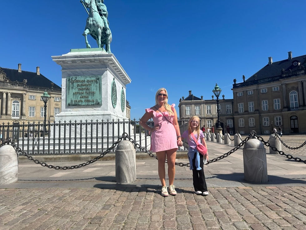 A woman and a girl in pink dresses stand in a sunlit cobblestone square beside an equestrian statue with historic buildings behind them.