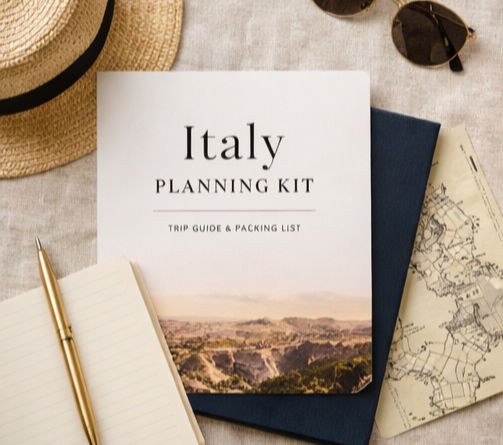 Italy planning kit with a landscape cover on a table, surrounded by a straw hat, sunglasses, pen, notebook, and a map. Neutral tones.