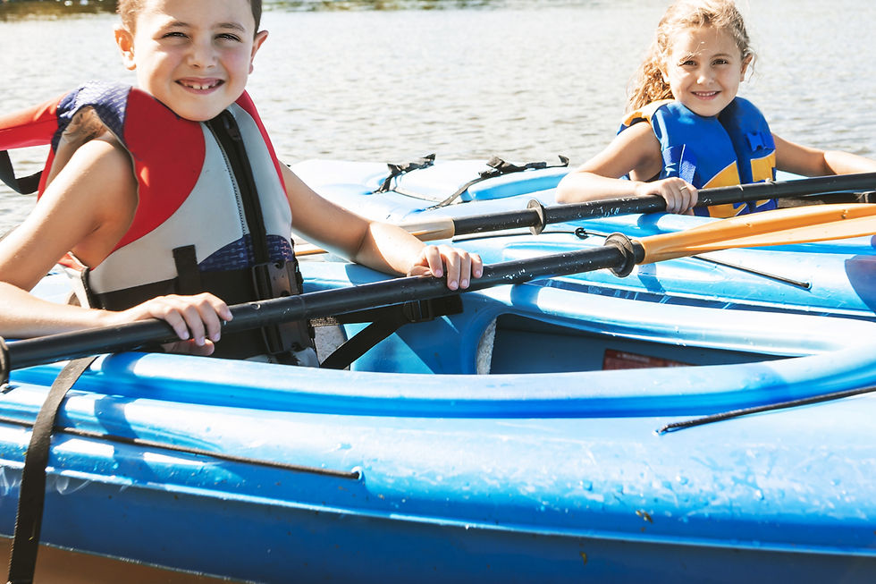 Kids in life jackets smile while sitting in blue kayaks on a sunny day, holding paddles, with water in the background.