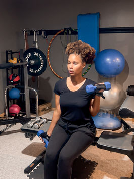 Woman in black outfit lifting blue dumbbells, seated in a home gym with exercise equipment and blue exercise ball in background. Focused mood.