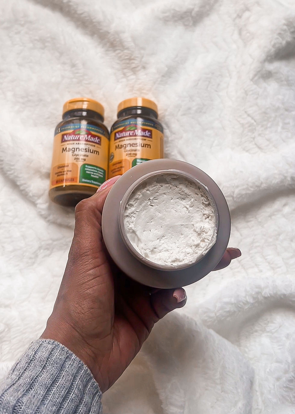 Hand holds open jar of white cream in front of two Nature Made Magnesium bottles on a fluffy white blanket.