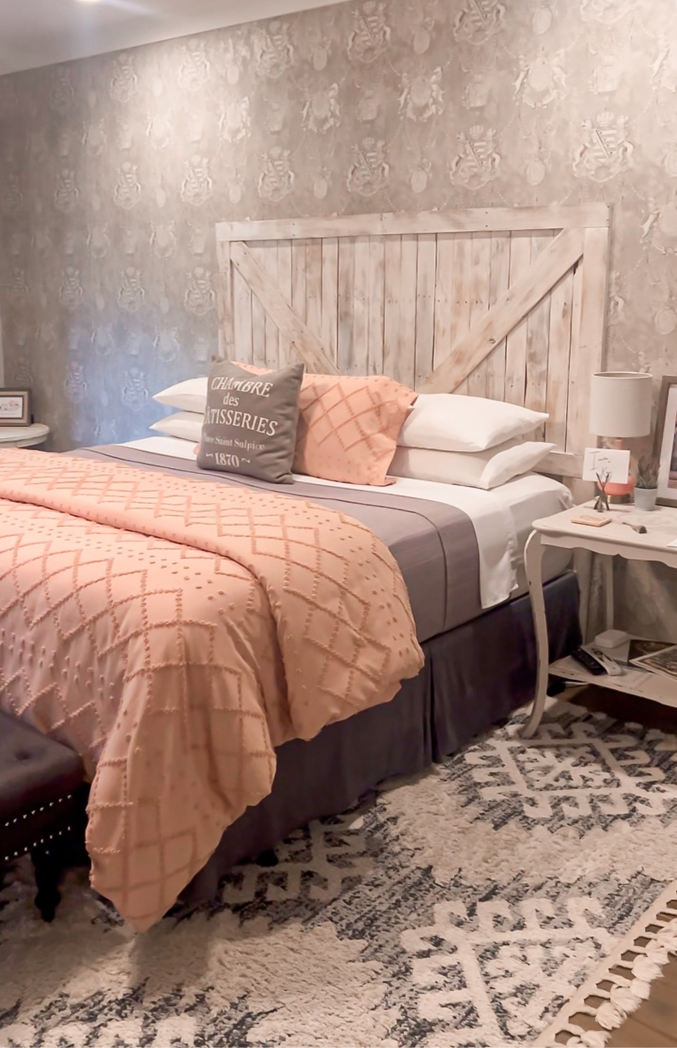 Cozy bedroom with rustic wood headboard, peach bedding, patterned wallpaper, bedside table, and pillow reading CHAMBRE des TISSERIES.