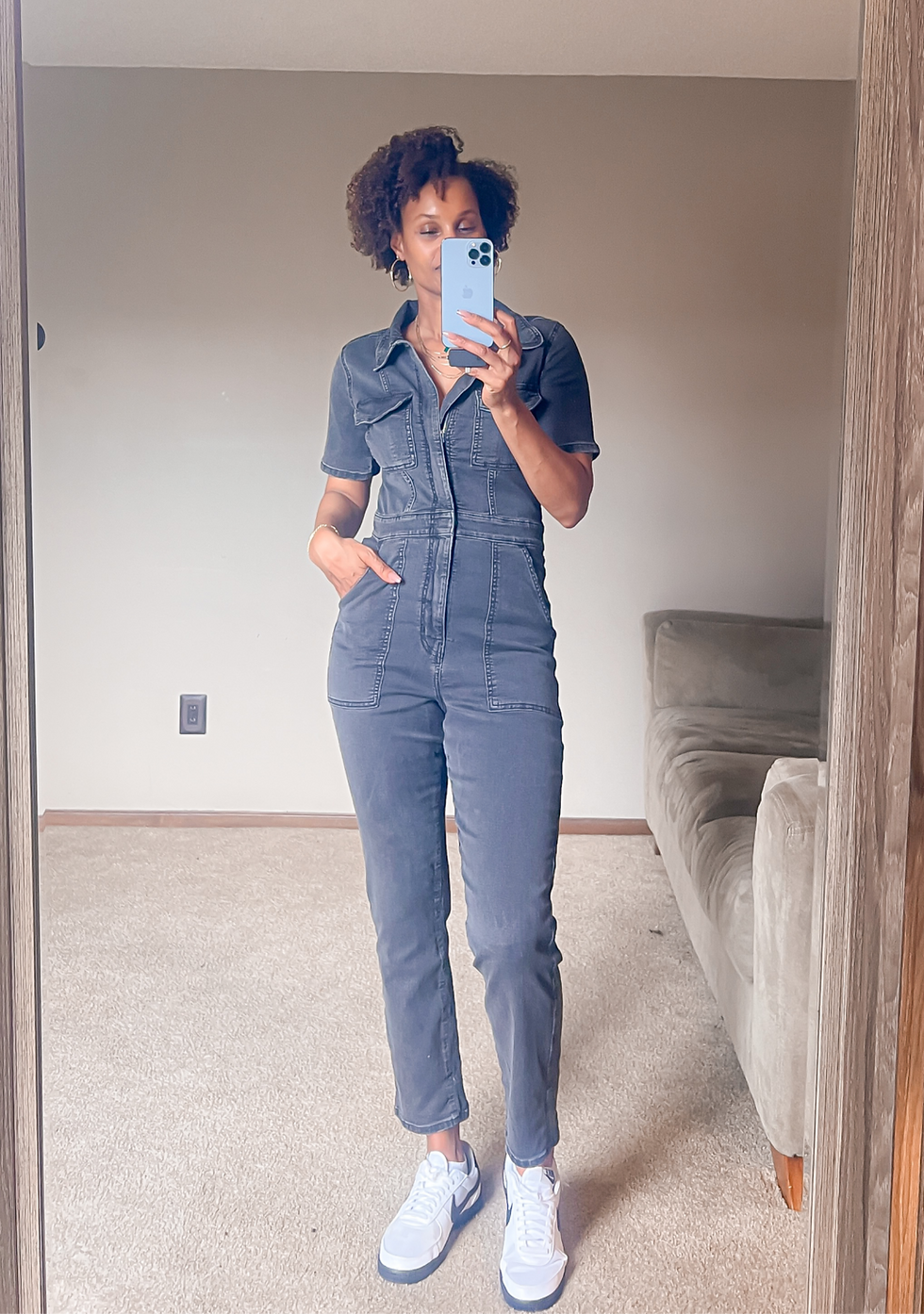 Woman takes a mirror selfie in a living room, wearing a gray denim jumpsuit and white sneakers, with a beige couch beside her.