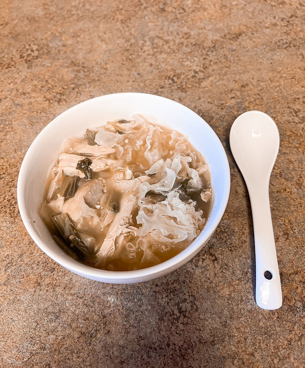 A bowl of soup with vegetables and noodles next to a white spoon on a brown countertop. The mood is calm and inviting.