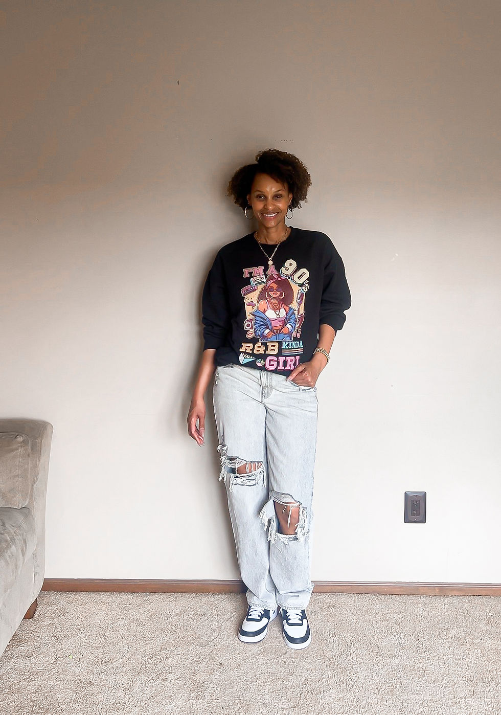 Smiling woman indoors against a blank wall, wearing a black graphic sweatshirt reading I’M A 90S R&B KINDA GIRL and ripped jeans.