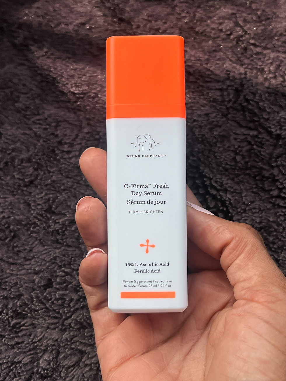 Hand holding a Drunk Elephant C-Firma Fresh Day Serum bottle with orange cap against a dark fuzzy background.