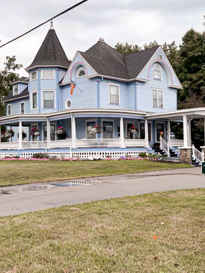 Victorian-style house with a blue exterior and turret, surrounded by a wraparound porch and flower baskets. Overcast sky, trees in background.