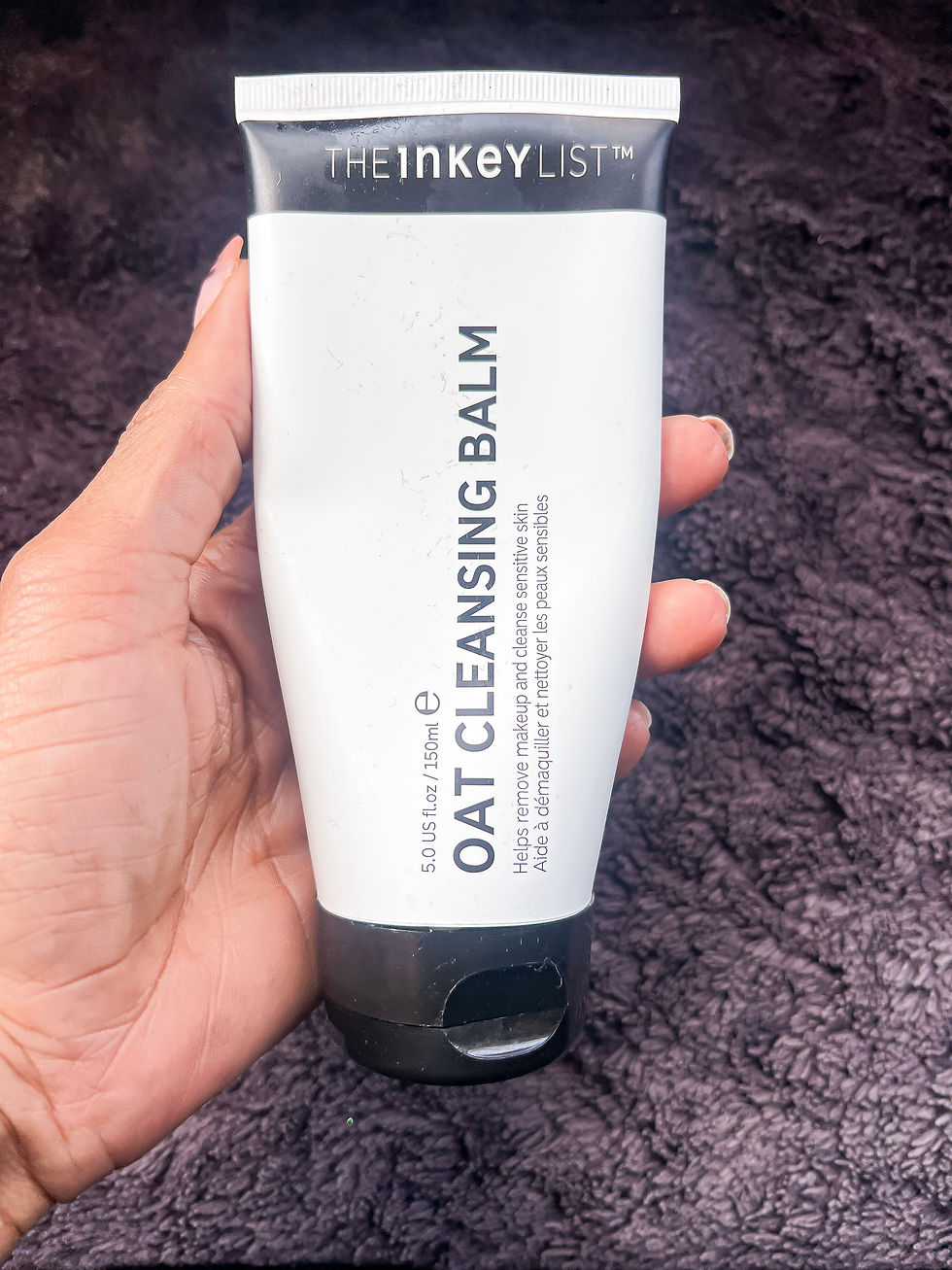 Hand holding The INKEY List Oat Cleansing Balm tube against a dark textured towel background.