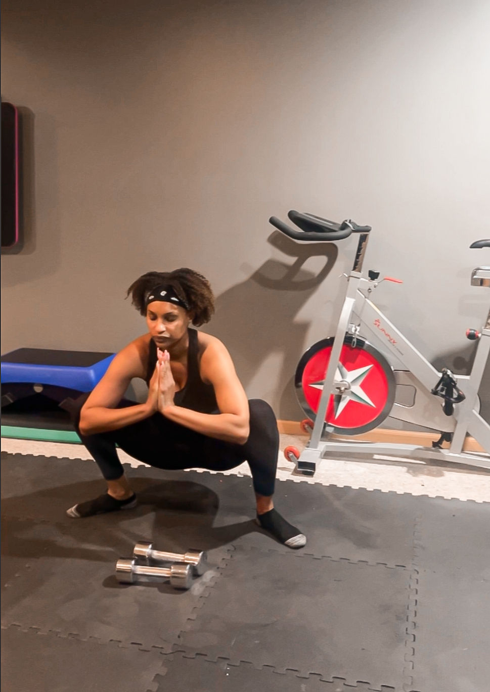 Woman squatting in a workout room, hands together in a prayer position. Exercise bike and dumbbells visible. Focused mood.