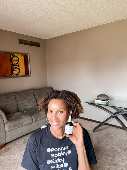 Smiling woman in a living room holds a skincare dropper bottle; black shirt reads Ronnie Bobby Ricky Mike.