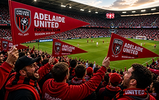 Passionate football supporters dressed in red fill a packed stadium, waving bright red club pennants with team branding while cheering toward the pitch, where a match unfolds under daylight with a vibrant crowd atmosphere.