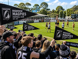 Supporters at a local Australian rules football match on a bright sunny day stand along the sideline waving black club pennants, cheering as players contest the ball on a green oval with goalposts, clubhouse, and scoreboard in the background.