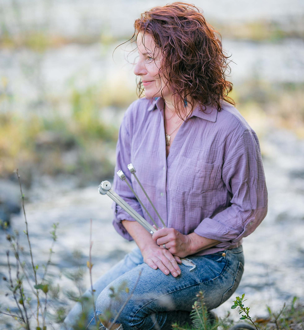 A woman, Noémi Kiss, wearing a purple button up shirt and jeans, kneeling by a river holding tuning forks