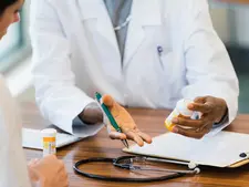 The image shows a doctor in a white coat, holding a prescription bottle in one hand and a pen in the other, sitting at a desk with a patient. A stethoscope, another prescription bottle, and a clipboard with papers are on the desk, indicating a medical consultation. The setting suggests a discussion about medication or treatment, likely in a clinical or hospital environment.