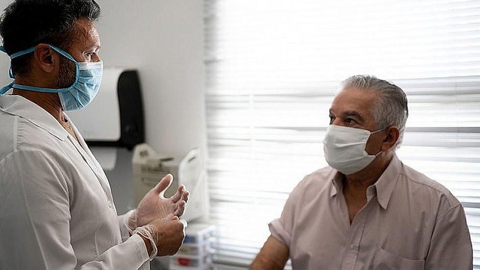 A doctor in a mask consults an elderly man in a bright medical office. Both wear white shirts and masks, with sunlight filtering through blinds.