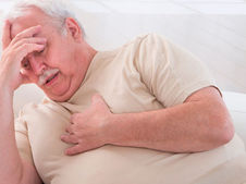 Elderly man in beige shirt clutching chest, looking in discomfort. He rests his head on his hand. Bright, minimal background.