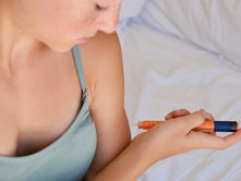 Person in a gray tank top injects themselves with an orange syringe. White bedding in the background, focused expression.