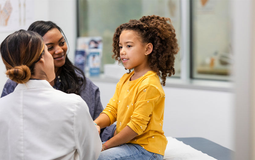 A young girl in a yellow sweater sits on an exam table in a doctor's office, speaking to a healthcare worker, with her mother nearby.