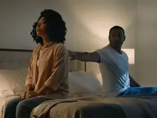 A person sits on a bed looking away, while another reaches out to them in a dimly lit bedroom, creating a tense mood.