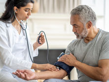 Doctor checks man's blood pressure in a bright room. She uses a stethoscope and cuff, appearing focused. The man looks relaxed.