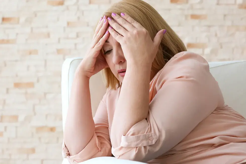 A woman with closed eyes holds her head in frustration, seated on a white couch. She's wearing a peach blouse, with a brick wall background.