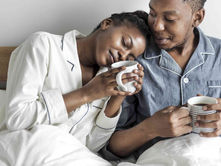 Couple in pajamas cuddling in bed, holding mugs. She leans on his shoulder, eyes closed, serene. White and blue pajamas, cozy mood.
