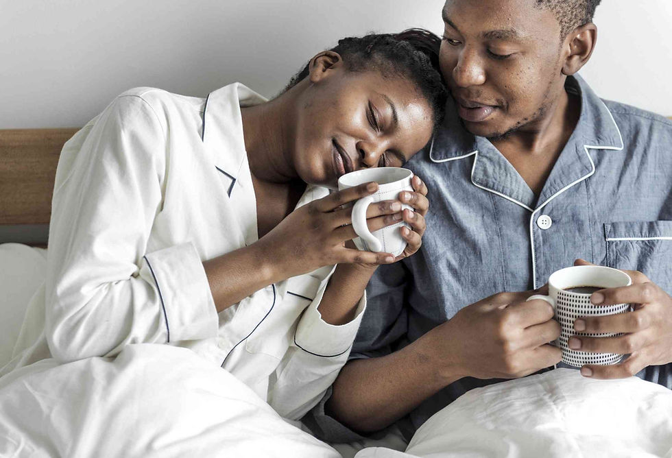 Couple in pajamas cuddling in bed, holding mugs. She leans on his shoulder, eyes closed, serene. White and blue pajamas, cozy mood.