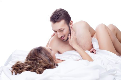A couple lying in bed under white sheets, gazing at each other affectionately with soft smiles. The background is bright and white.