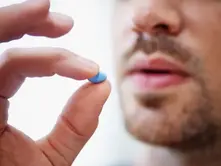 A person holds a small blue pill between fingers near their lips, close-up. The background is blurred, focusing on the pill and fingers.