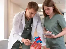 Two medical professionals examine a model of a heart in a bright, clinical setting. One takes notes, both appear focused.