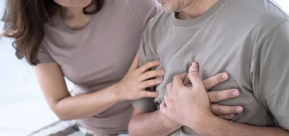 Woman reassuring man clutching chest, both in gray tops, expressing concern. Indoor setting with a bright background, conveying urgency.