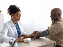 Doctor in a lab coat checks an elderly man's blood pressure in a bright, clinical office. The mood is calm and focused.