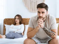 Man with tattoo sits pensively on bed, while woman sits in background looking away. Bright bedroom, neutral colors, tense mood.